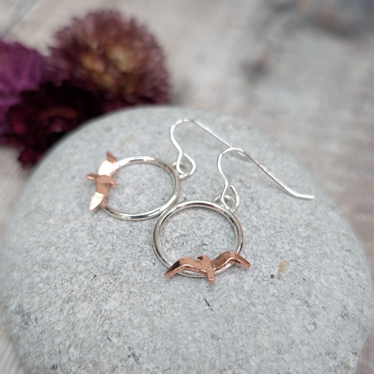 Silver Circle Earrings with Copper Birds resting on a smooth grey stone, each earring featuring a hand-cut copper bird perched along the bottom of the silver circle, with blurred dried purple flowers in the background.