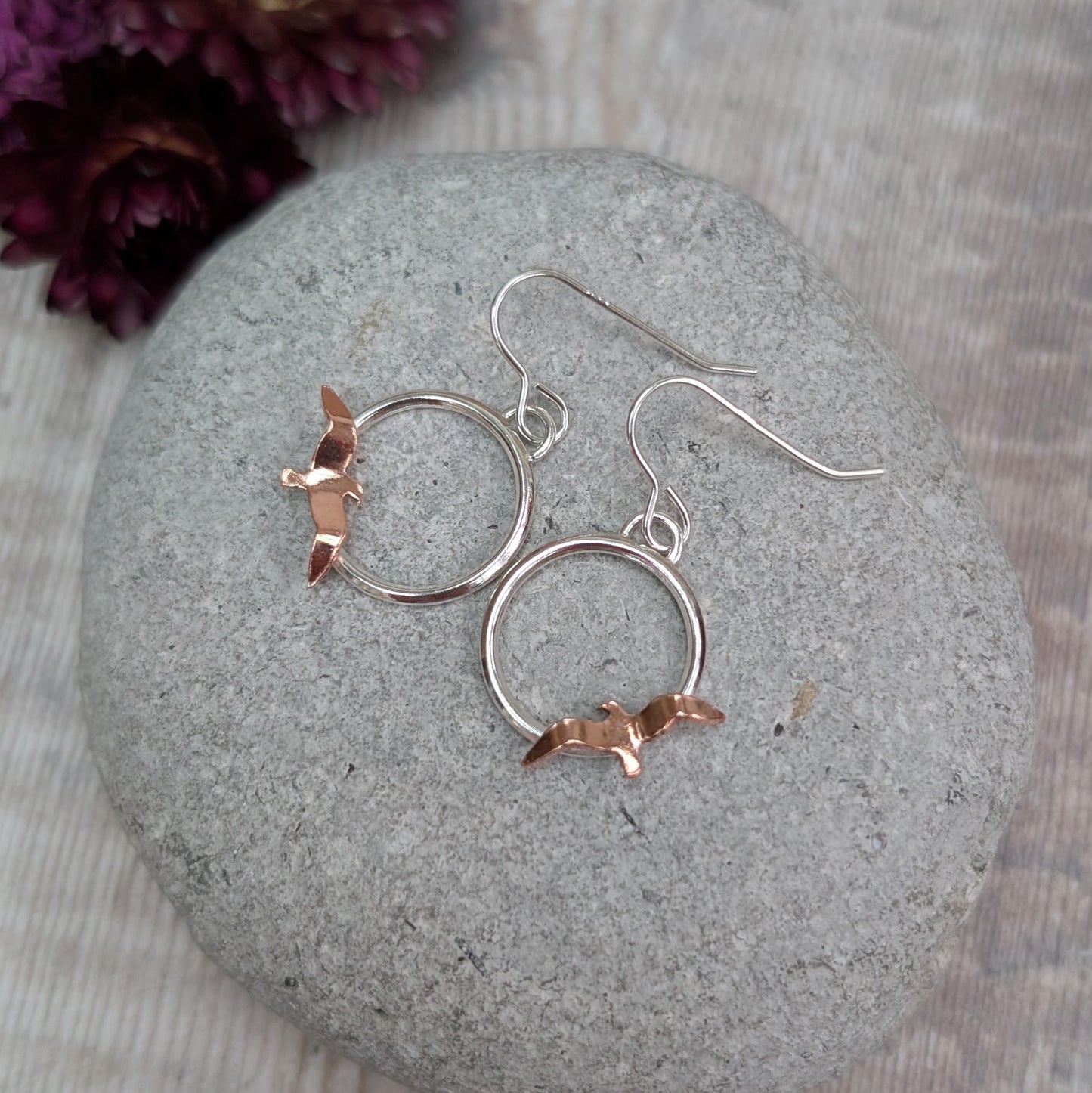 A top-down view of Silver Circle Earrings with Copper Birds displayed on a rounded grey stone, showing the simple silver hooks and hand-cut copper birds against a light wooden background.