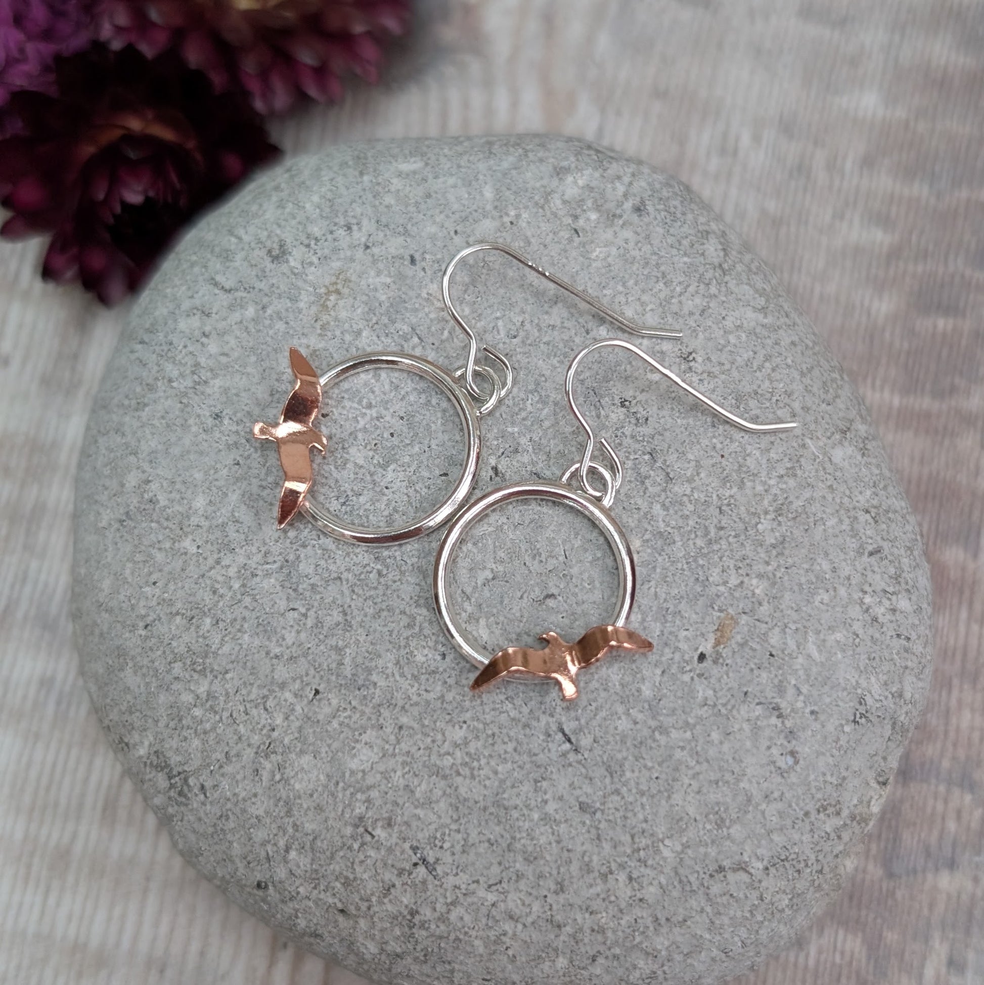 A top-down view of Silver Circle Earrings with Copper Birds displayed on a rounded grey stone, showing the simple silver hooks and hand-cut copper birds against a light wooden background.
