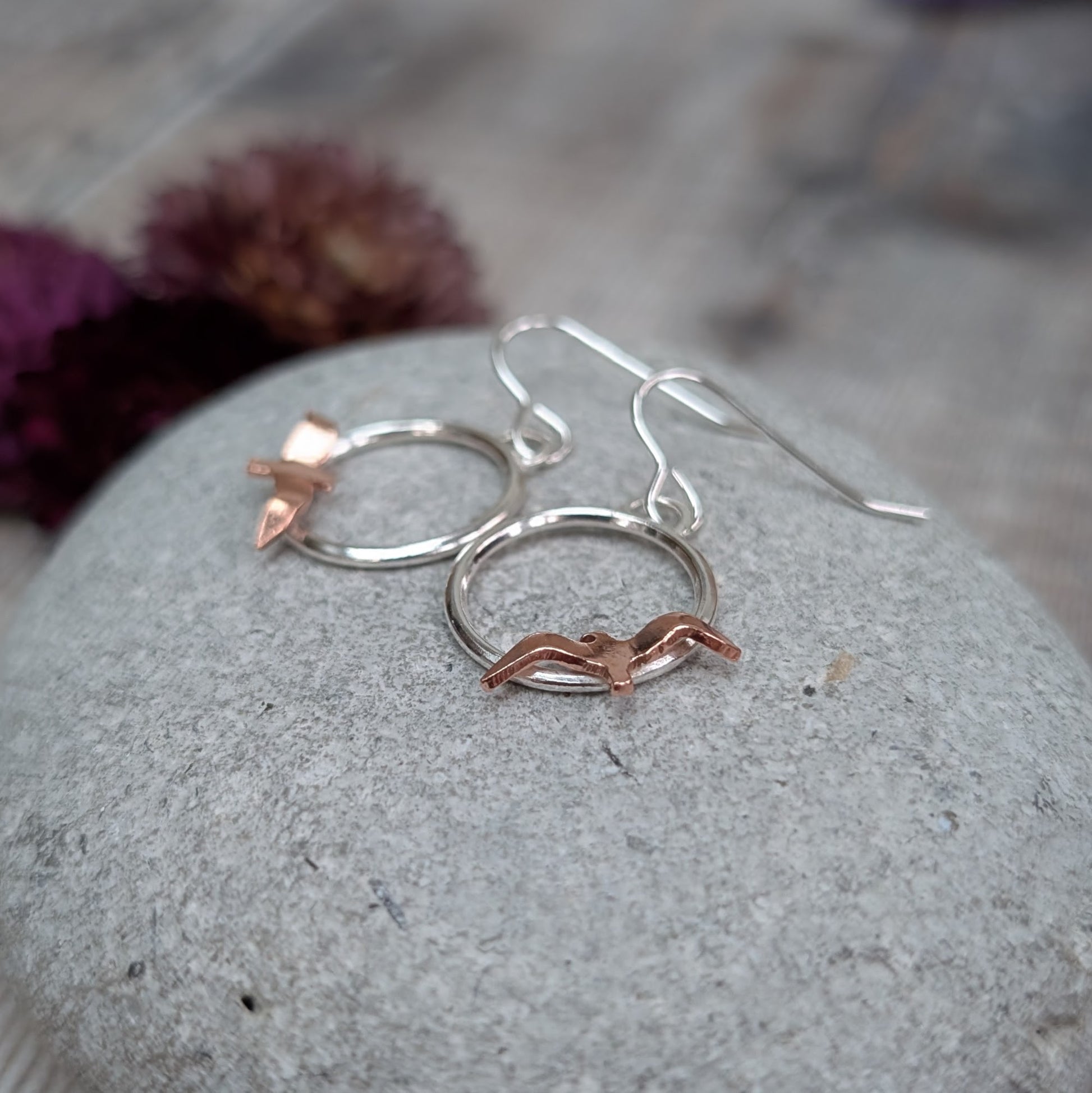 An angled close-up of Silver Circle Earrings with Copper Birds placed on a grey stone, highlighting the warm copper birds sitting on the lower edge of each silver circle.