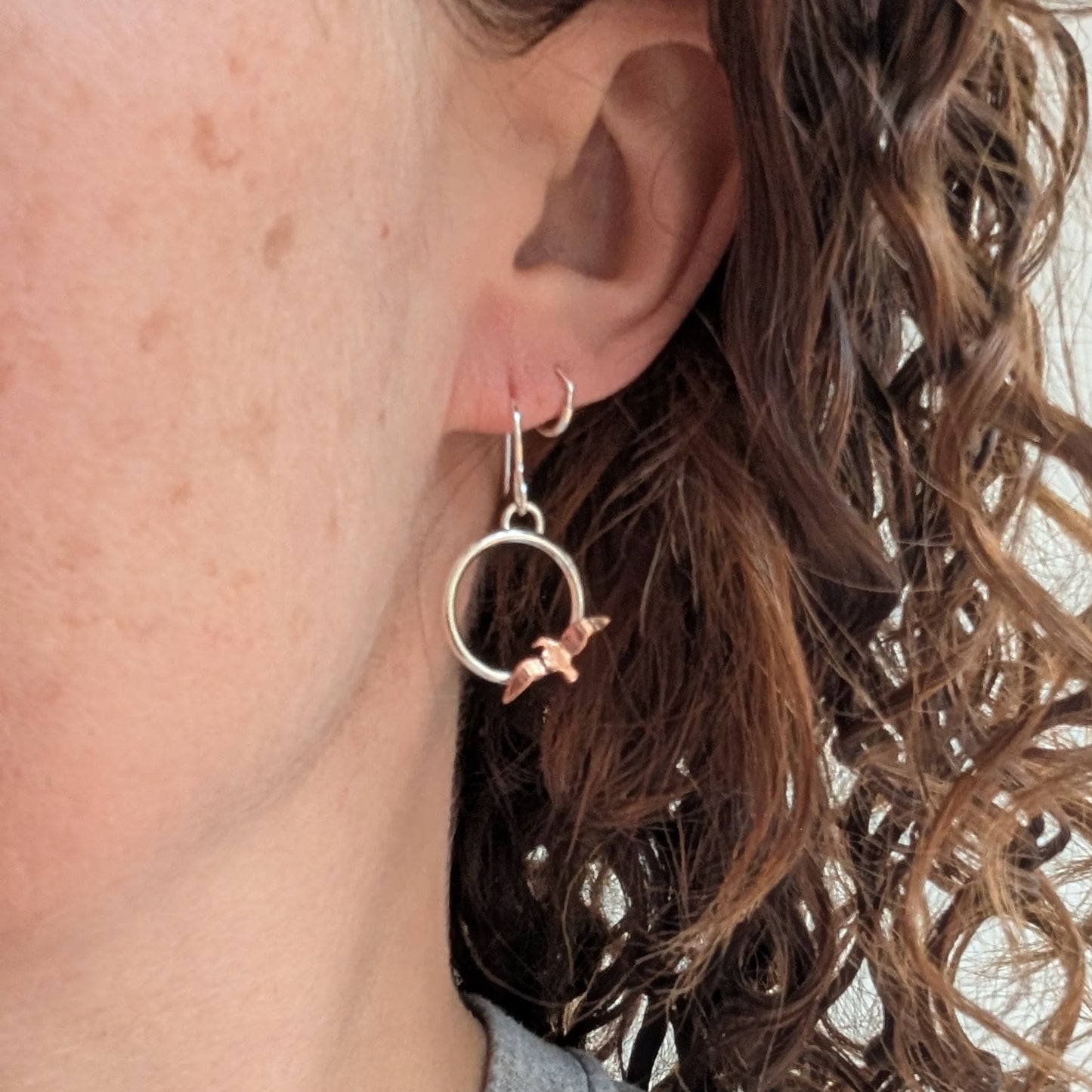 One of the Silver Circle Earrings with Copper Birds being worn on an ear with curly brown hair, showing the silver circle and hand-cut copper bird hanging lightly.