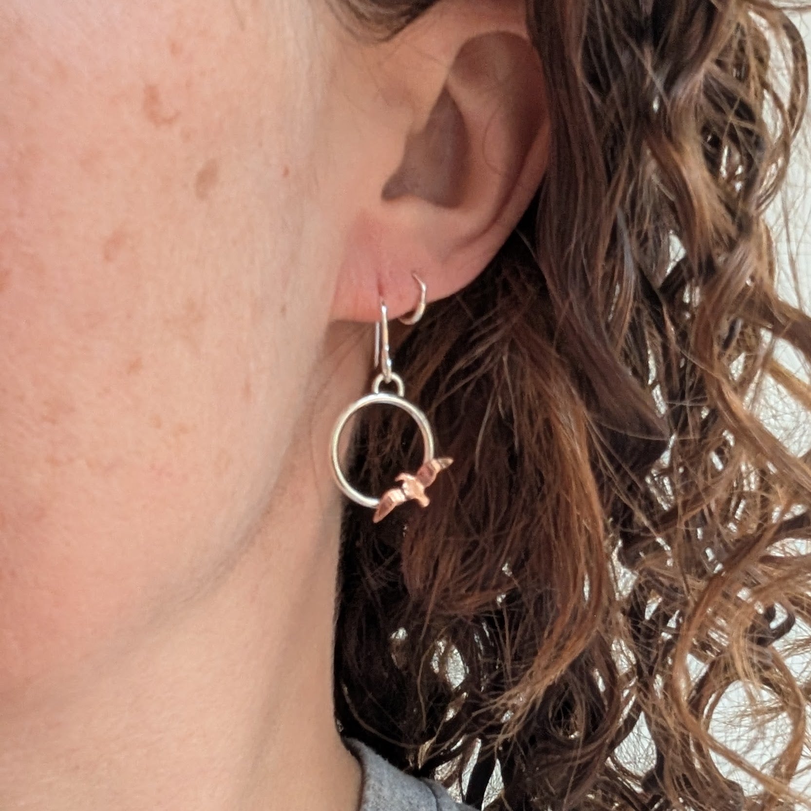 One of the Silver Circle Earrings with Copper Birds being worn on an ear with curly brown hair, showing the silver circle and hand-cut copper bird hanging lightly.