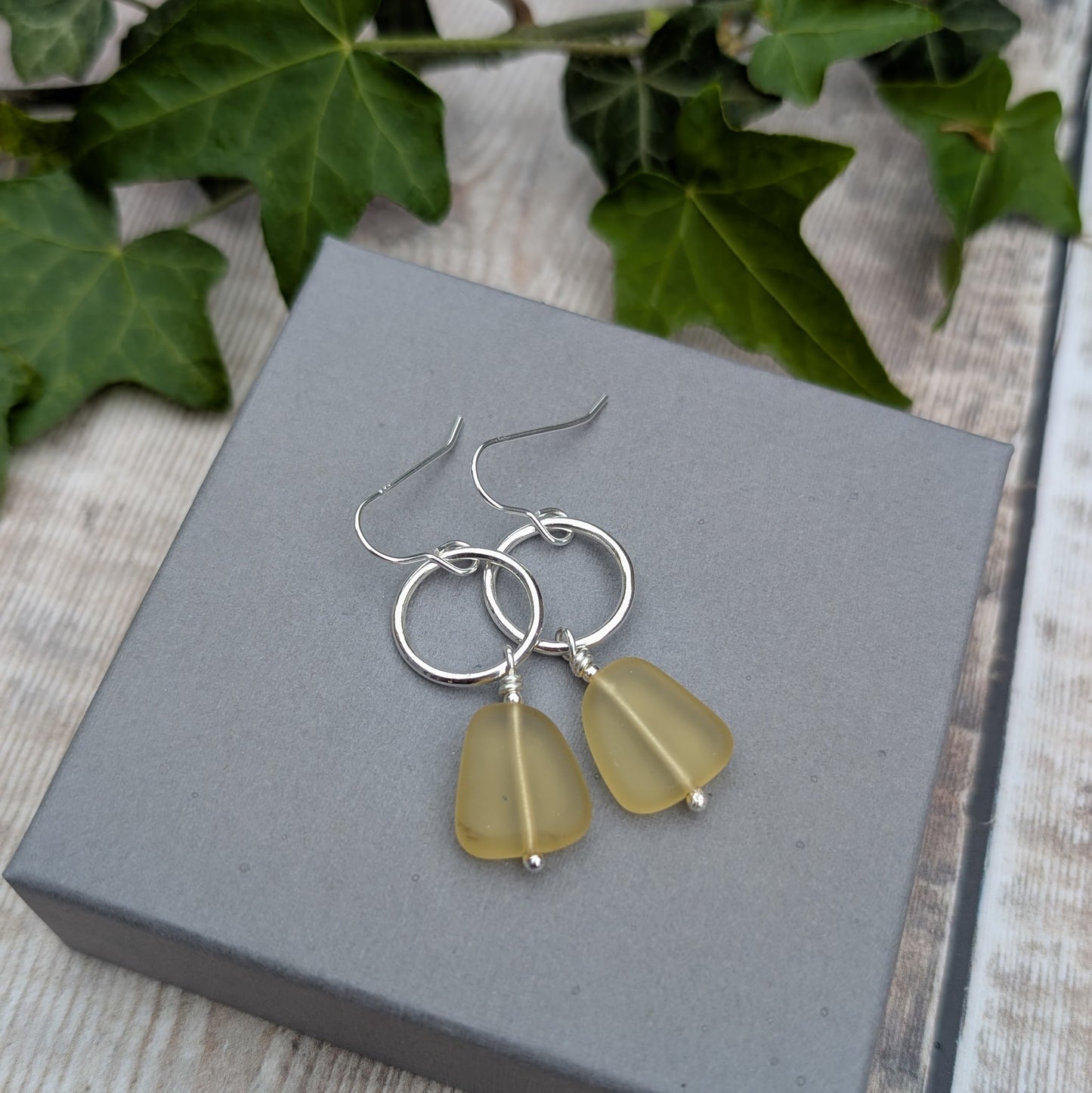 Silver Circle Earrings with Yellow Sea Glass Beads resting on a grey jewellery box with green ivy leaves in the background.