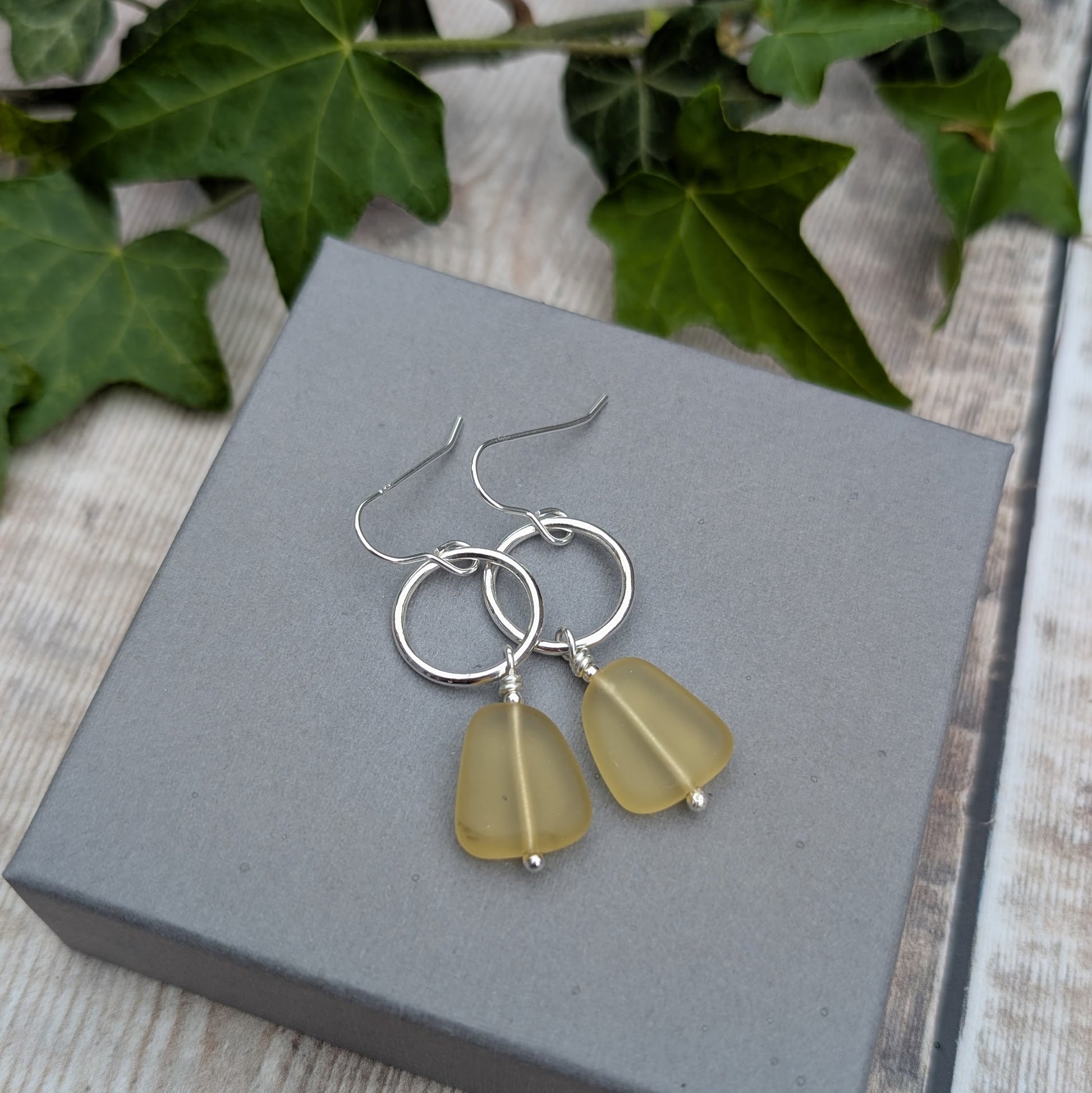 Silver Circle Earrings with Yellow Sea Glass Beads resting on a grey jewellery box with green ivy leaves in the background.