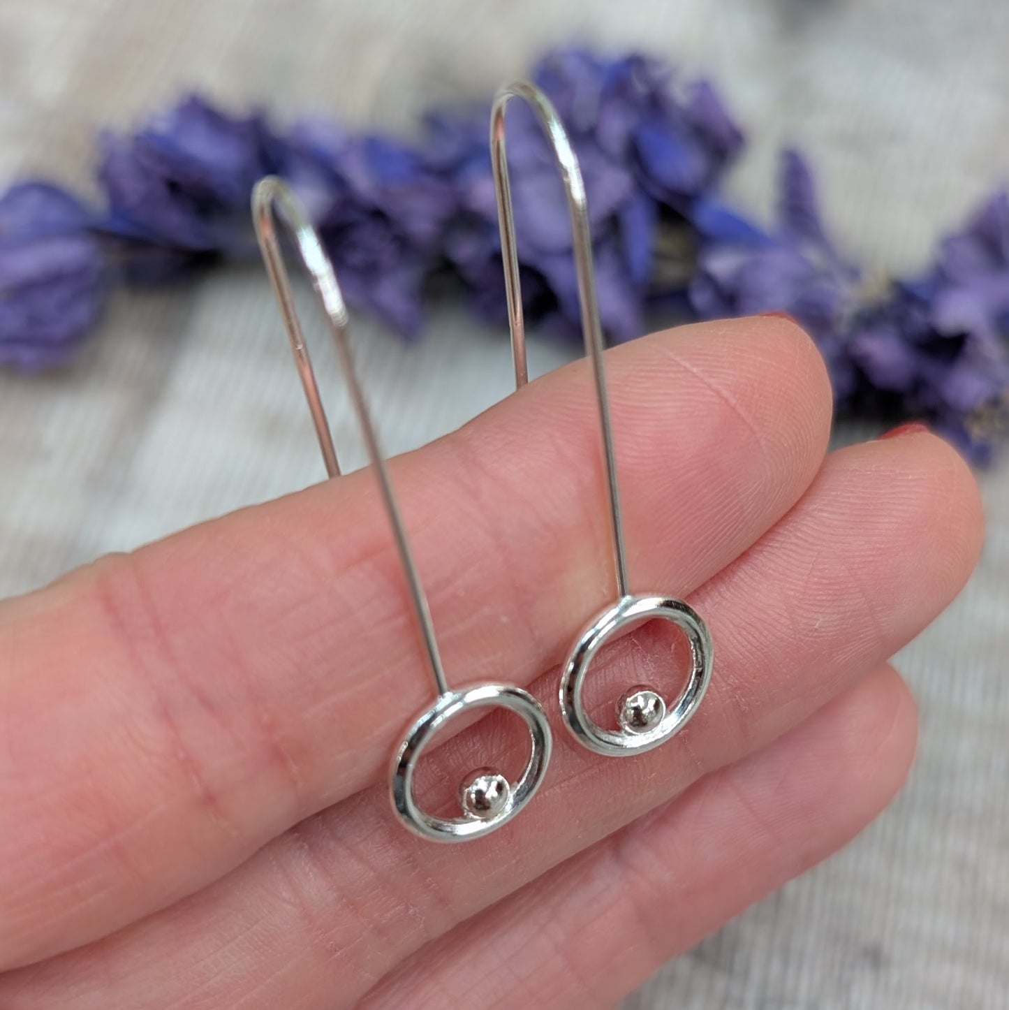 A close-up of a hand holding a pair of silver circle and dot long earrings, showing the polished silver circles with a small silver dot inside each one, attached to long ear wires.