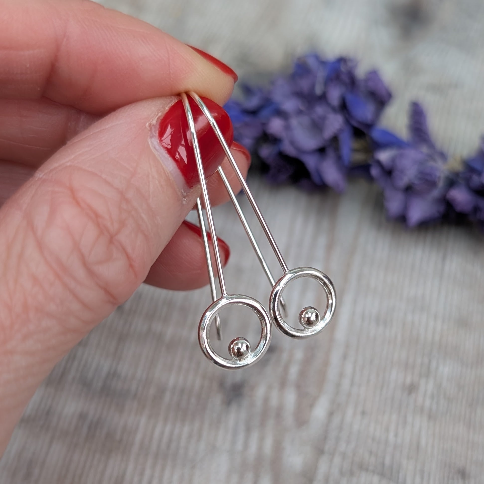 A hand with red nail polish holding the silver circle and dot long earrings, highlighting their sleek long wire design and shiny circular detail with a tiny dot inside each circle.