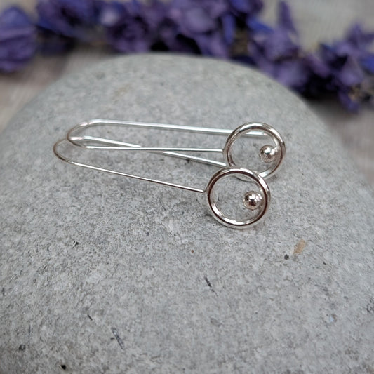The silver circle and dot long earrings resting on a smooth grey stone, showing their long, delicate ear wires and minimalist circular design with the small dot accent.