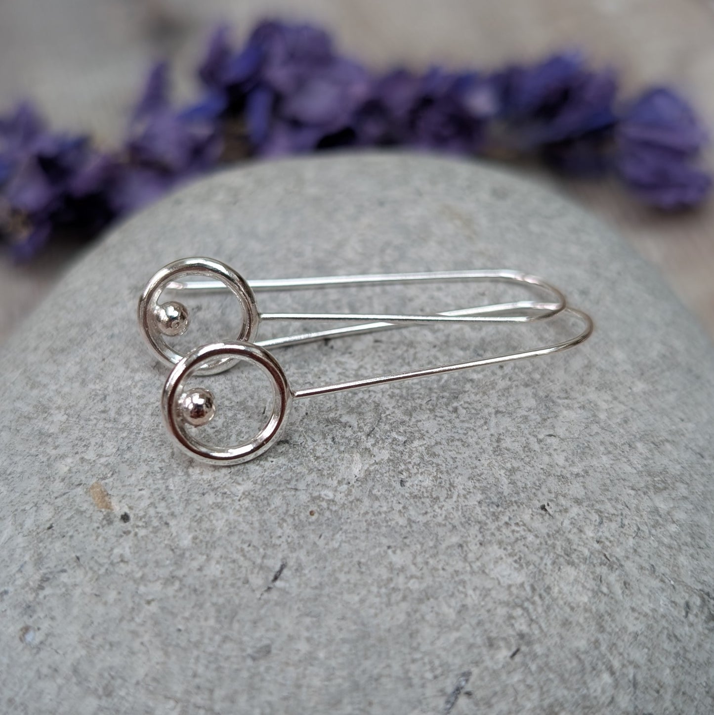 A side view of the silver circle and dot long earrings displayed on a grey stone, showing the long drop shape and the shiny silver circle with a small dot at the base of each one.
