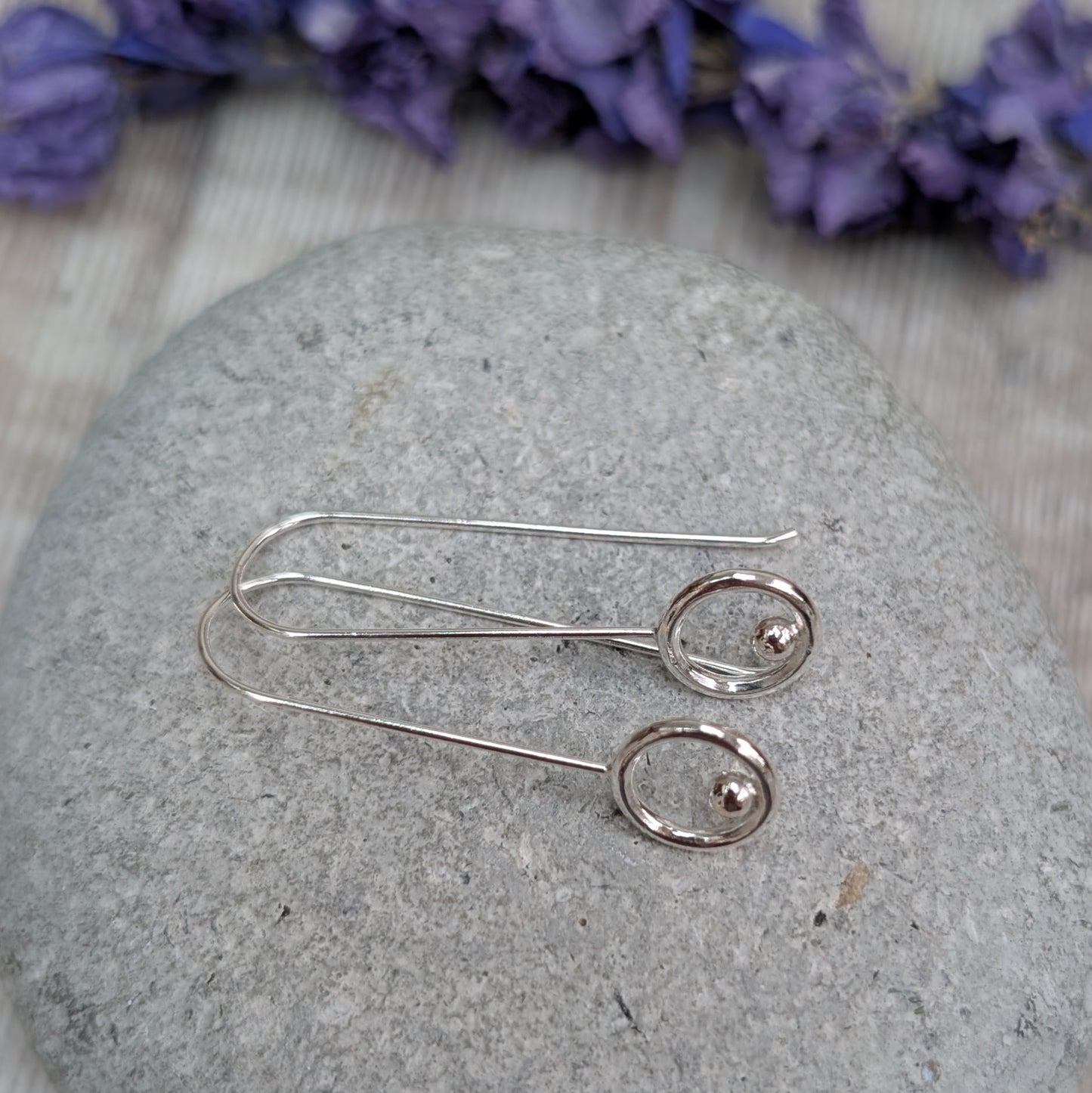 The silver circle and dot long earrings lying flat on a grey stone background, with the long wire hooks and polished circular details clearly visible.