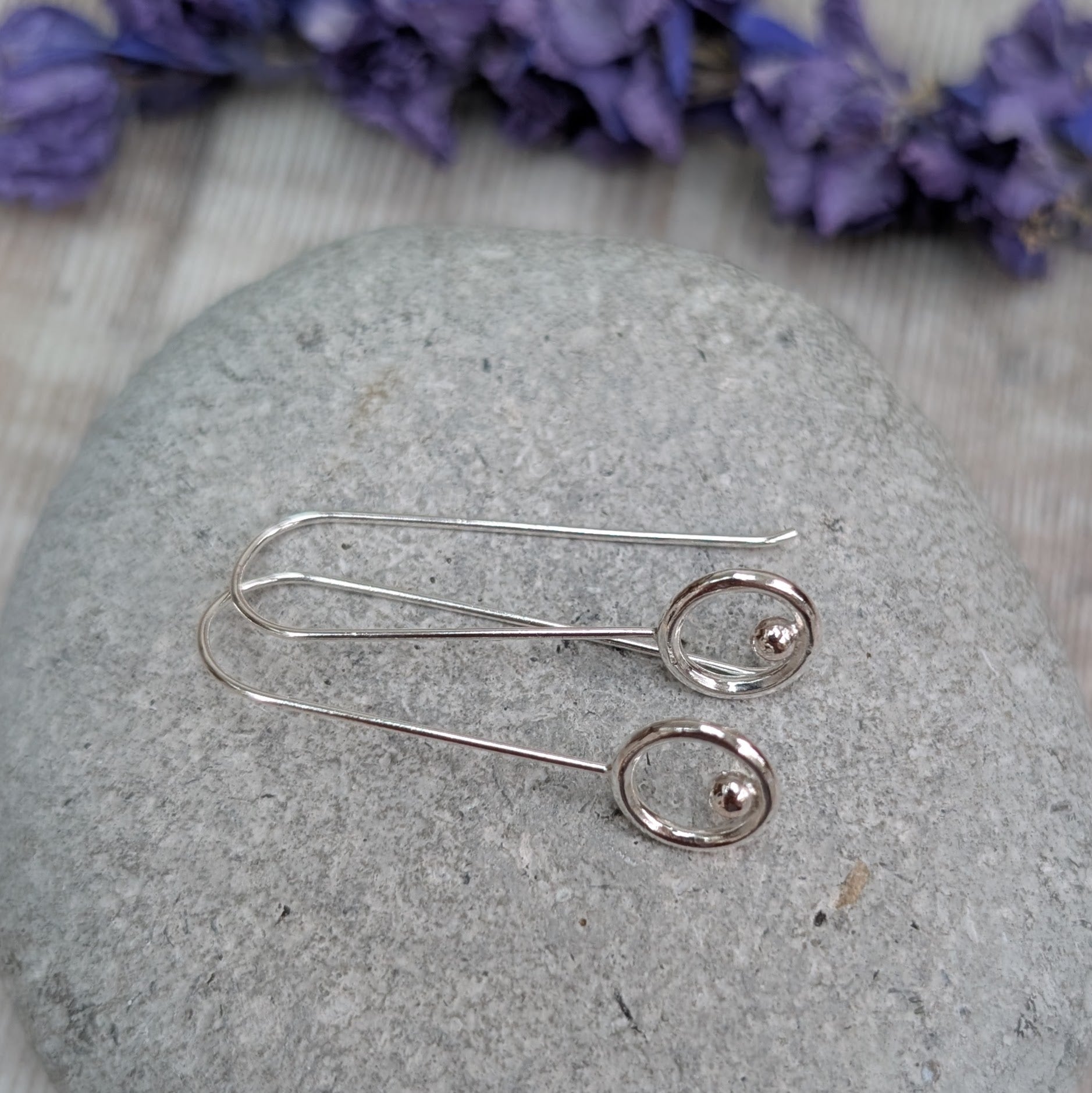 The silver circle and dot long earrings lying flat on a grey stone background, with the long wire hooks and polished circular details clearly visible.
