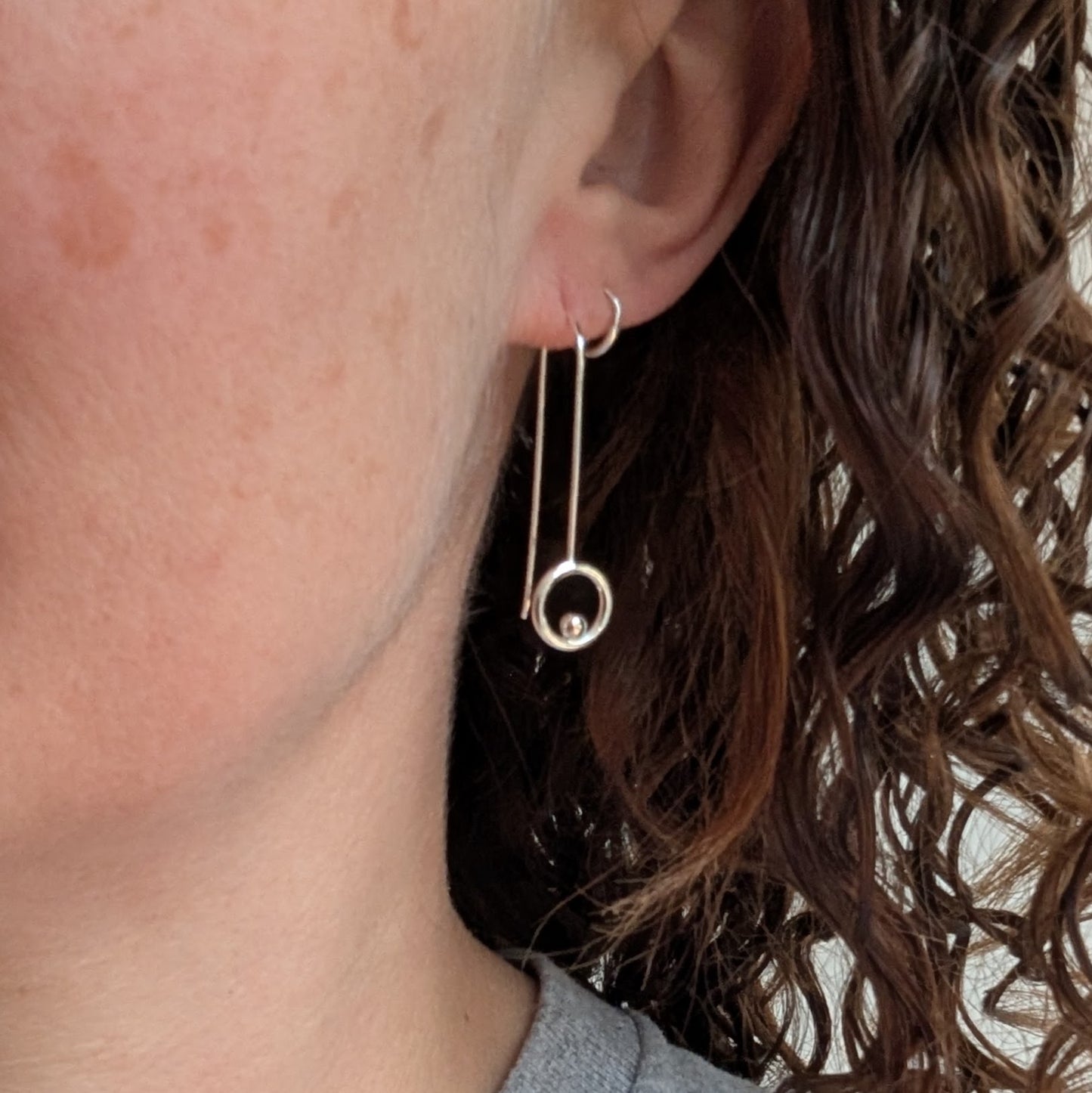 A close-up of a person wearing one of the silver circle and dot long earrings, showing how the long wire drop hangs neatly with the polished silver circle and dot resting just below the earlobe.