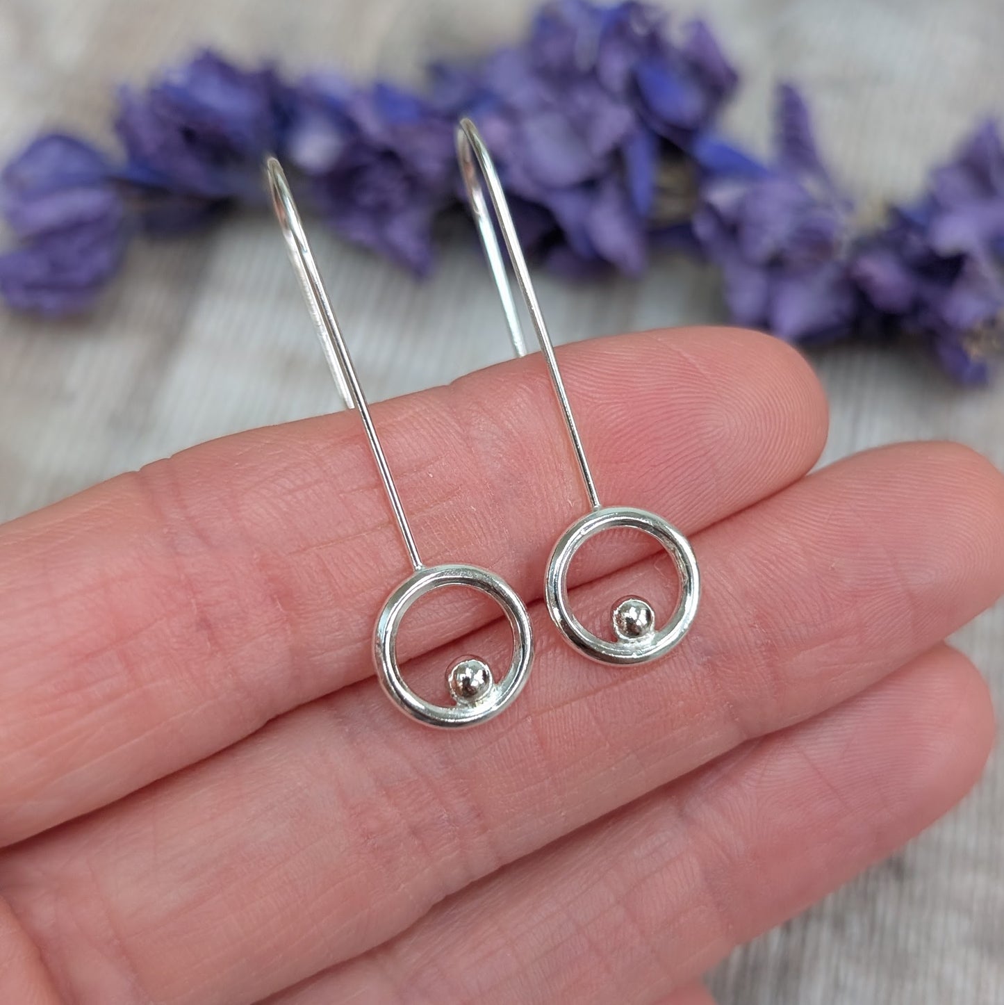 A close-up of a hand holding the silver circle and dot long earrings, showing the shiny silver circles with a tiny dot inside each one and the long, elegant ear wires.