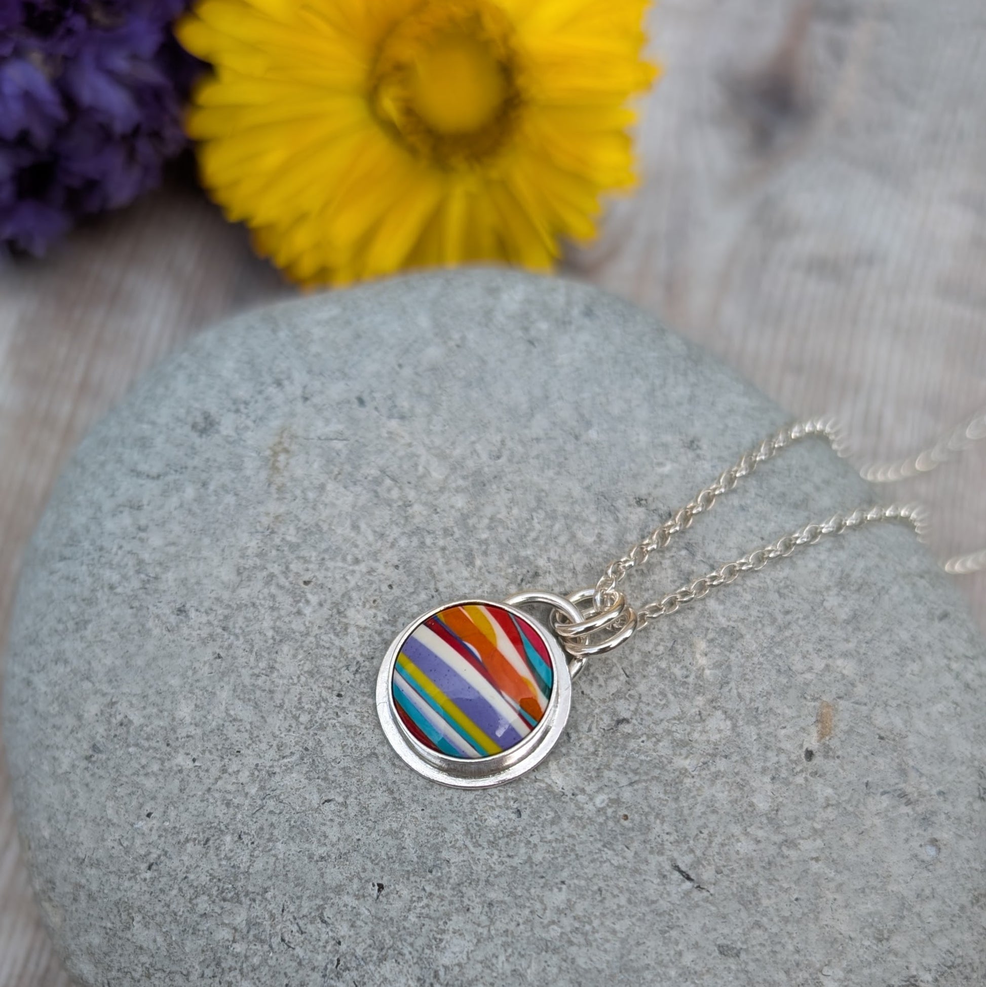 Silver Colourful Small Round Surfite Necklace resting on a grey stone with yellow and purple flowers in the background