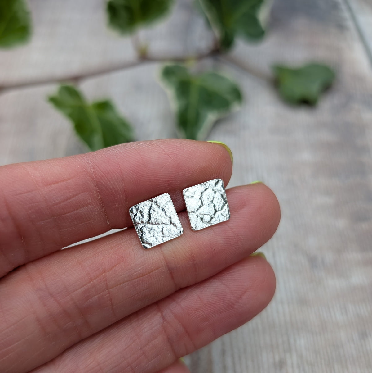 Silver cracked earth pattern square stud earrings held on fingertips, showing their small square shape and textured silver surface.