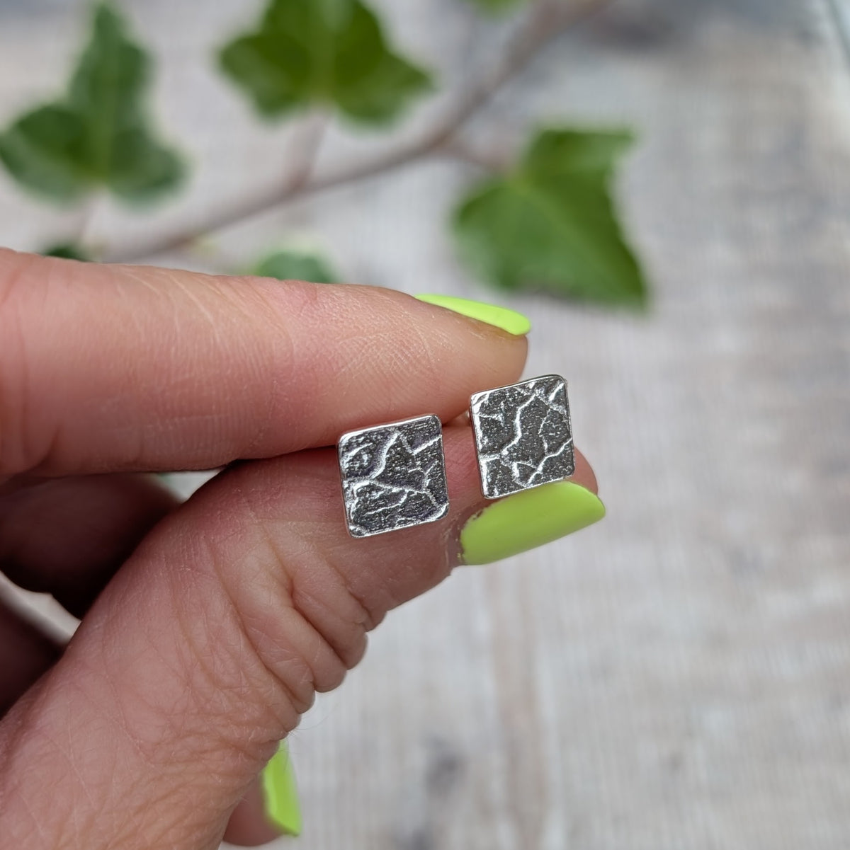Silver cracked earth pattern square stud earrings held between fingers, showing the textured pattern on the front of each earring.