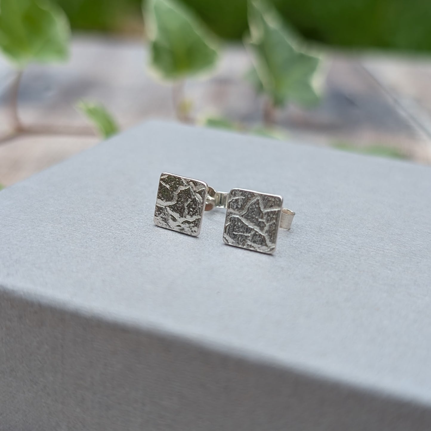 Silver cracked earth pattern square stud earrings displayed upright on a grey jewellery box with green ivy in the background.