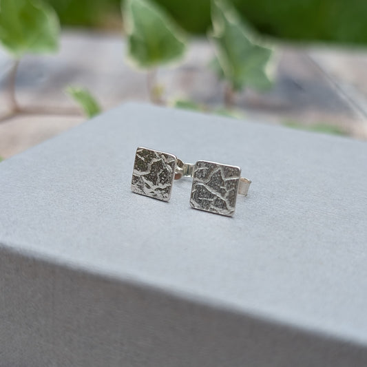 Silver cracked earth pattern square stud earrings displayed upright on a grey jewellery box with green ivy in the background.