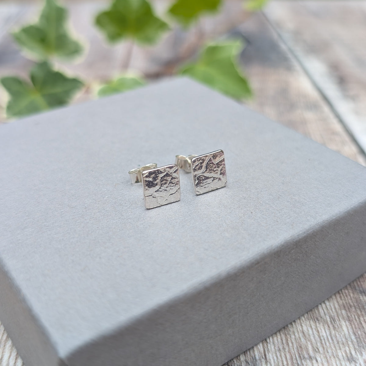 Pair of silver cracked earth pattern square stud earrings displayed on a grey box, showing the textured square fronts and stud fittings.