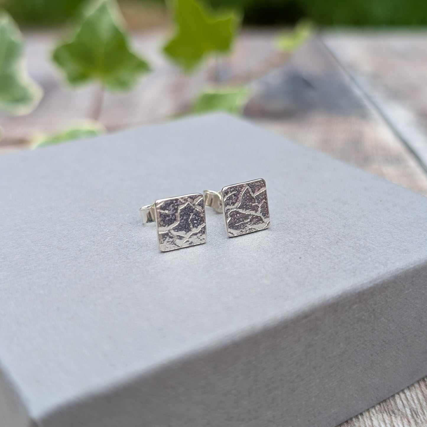 Close up of silver cracked earth pattern square stud earrings on a grey jewellery box, showing the detailed textured pattern.