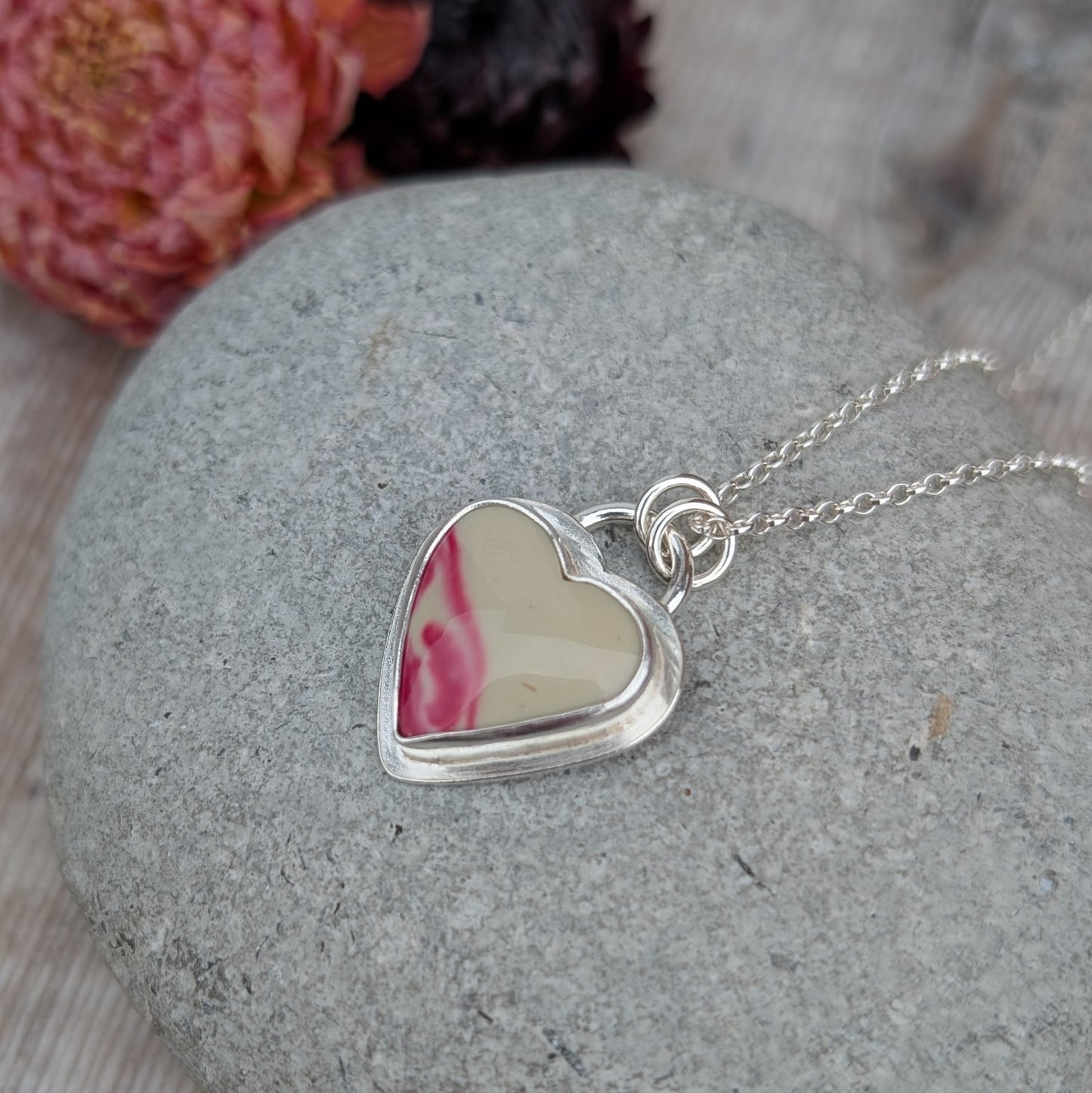 Silver heart necklace with cream and pink Surfite stone, shown on a grey pebble with flowers blurred in the background.