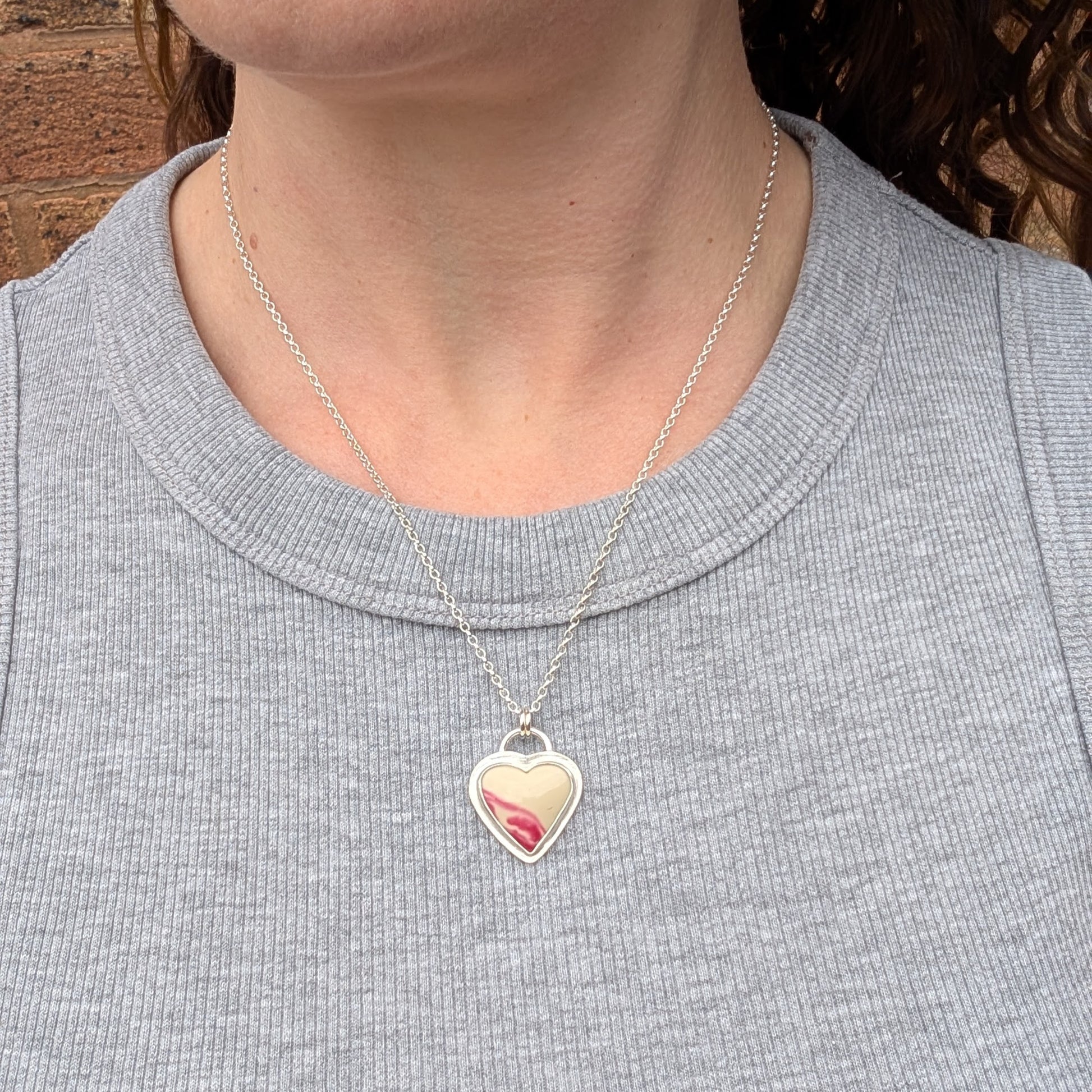 Silver heart necklace with cream and pink Surfite stone worn on a model, shown on a fine sterling silver chain against a grey ribbed top.