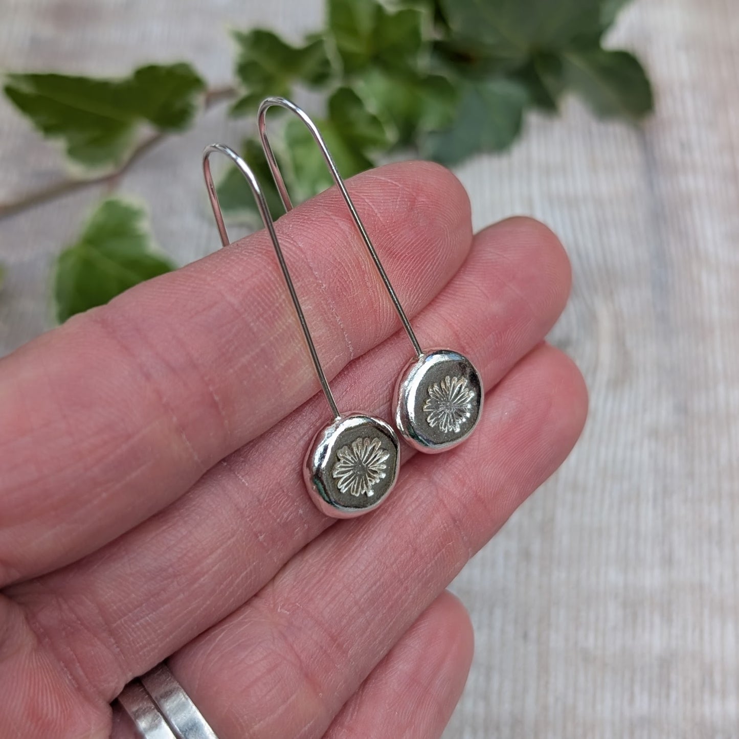 close up of silver daisy pebble long earrings on a hand, showing the daisy pattern stamped into each recycled Sterling Silver pebble.
