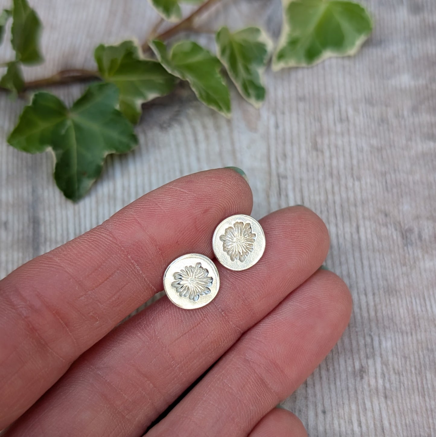 silver daisy stud earrings handmade from recycled Sterling Silver, held on fingertips with a leafy background.