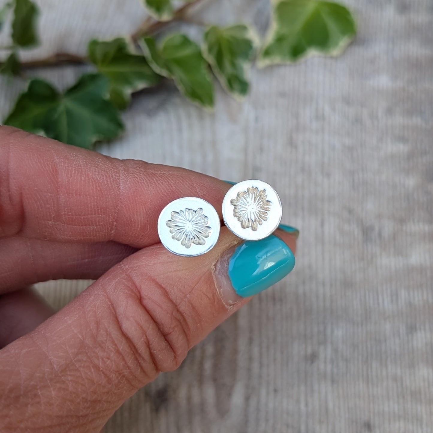 Close up of silver daisy stud earrings held between fingers, showing the hand stamped flower pattern on the round silver studs.
