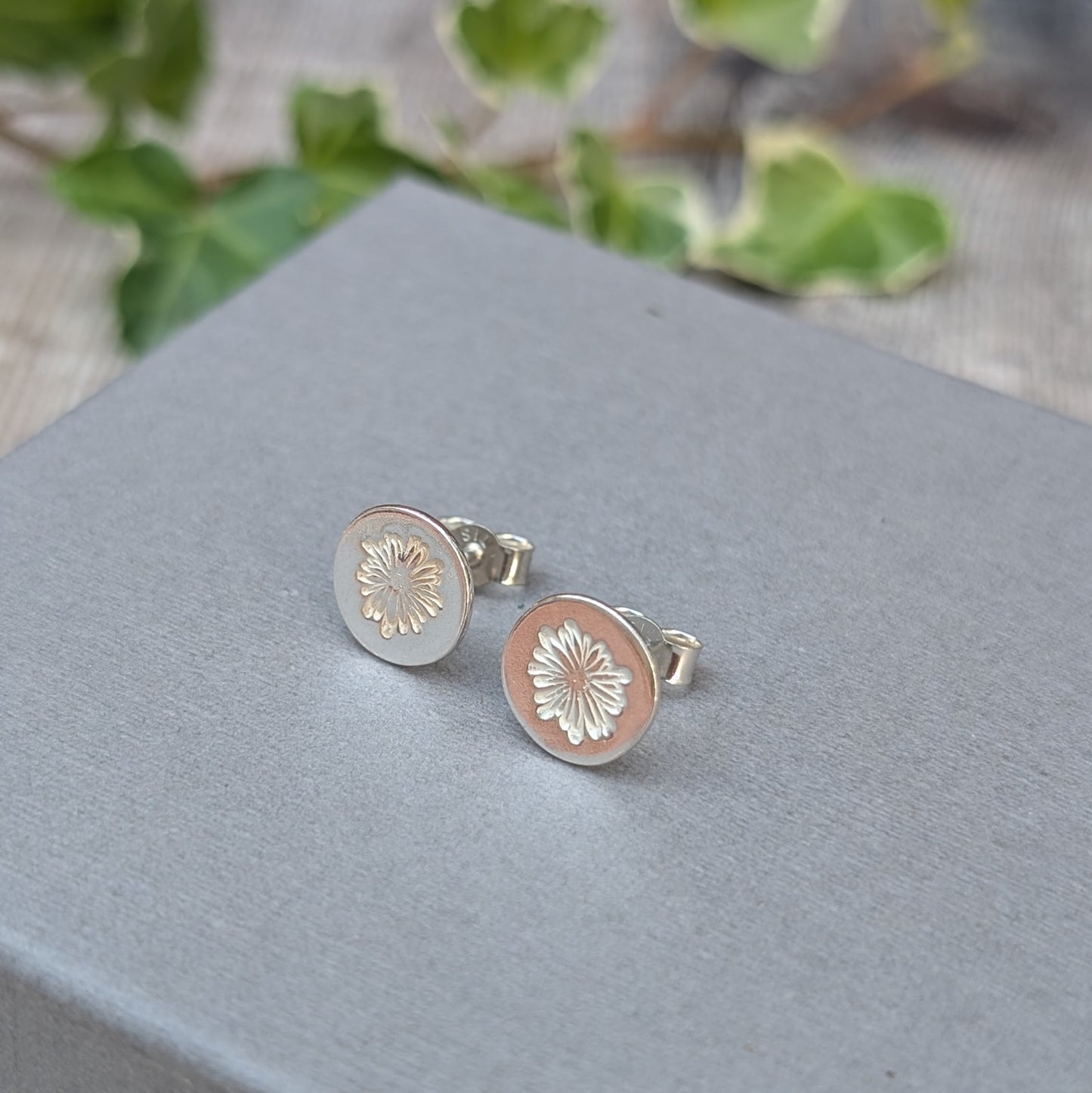 silver daisy stud earrings displayed on a grey jewellery box, showing the round shape, stamped daisy design and butterfly backs.