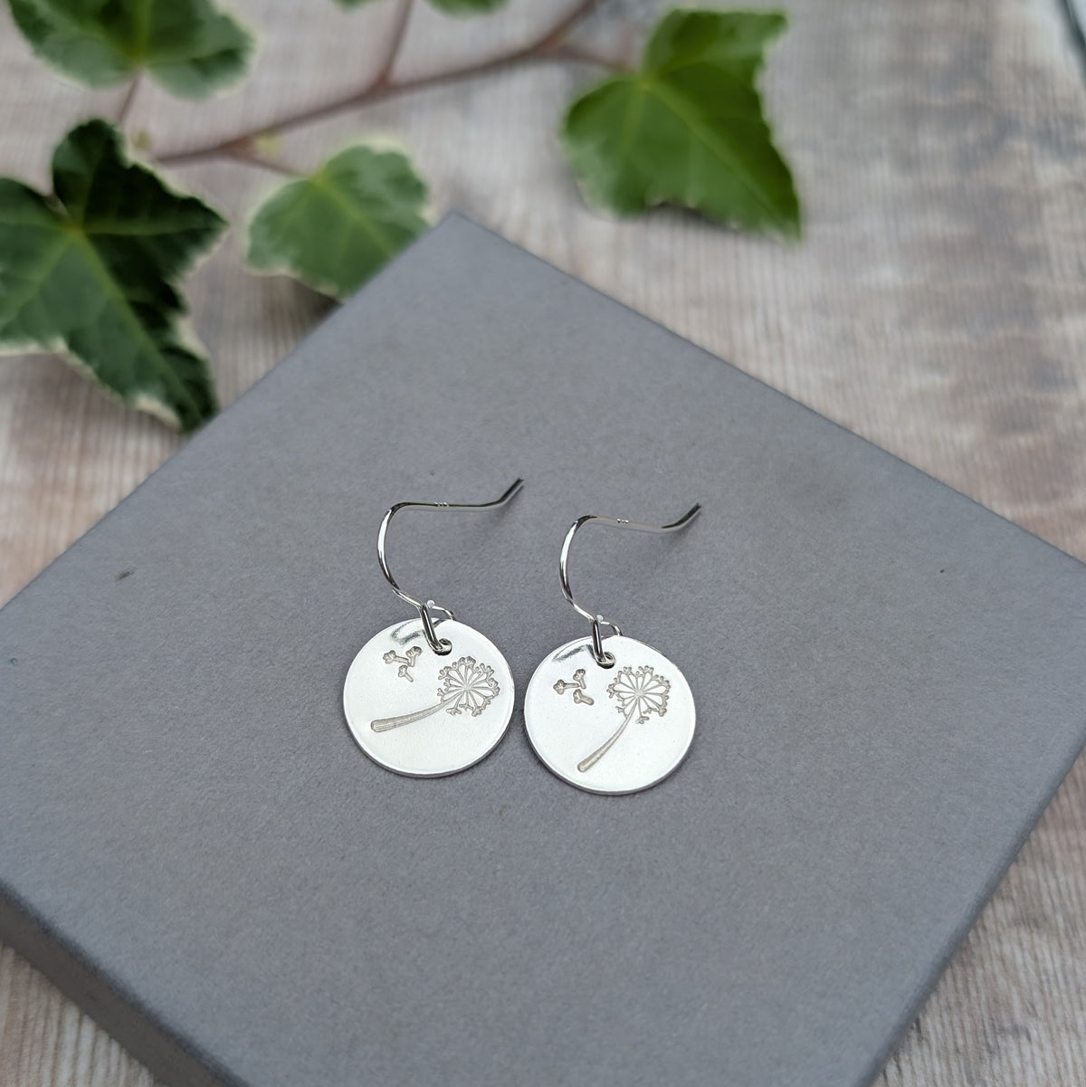Silver dandelion and wishes disc earrings displayed on a grey jewellery box with green leaves in the background.