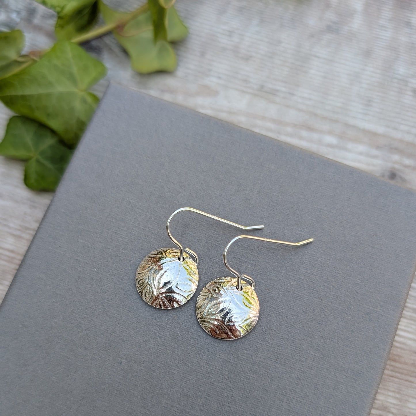 Silver Dome Disc Earrings with Leaf Pattern placed on a grey background with soft greenery in the corner.