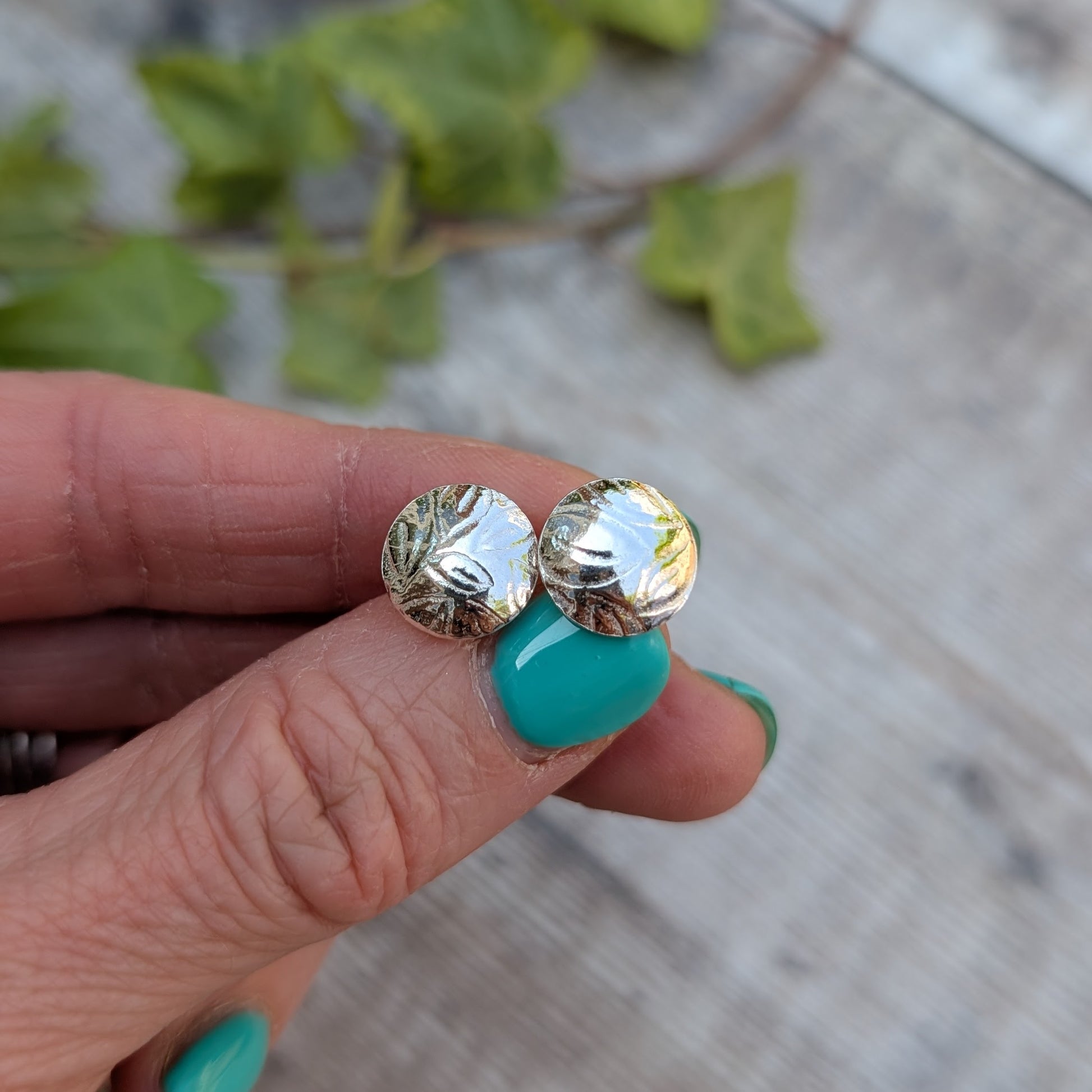 Silver Dome Disc Stud Earrings with Leaf Pattern held between fingers with greenery in the background, showing the handmade texture.