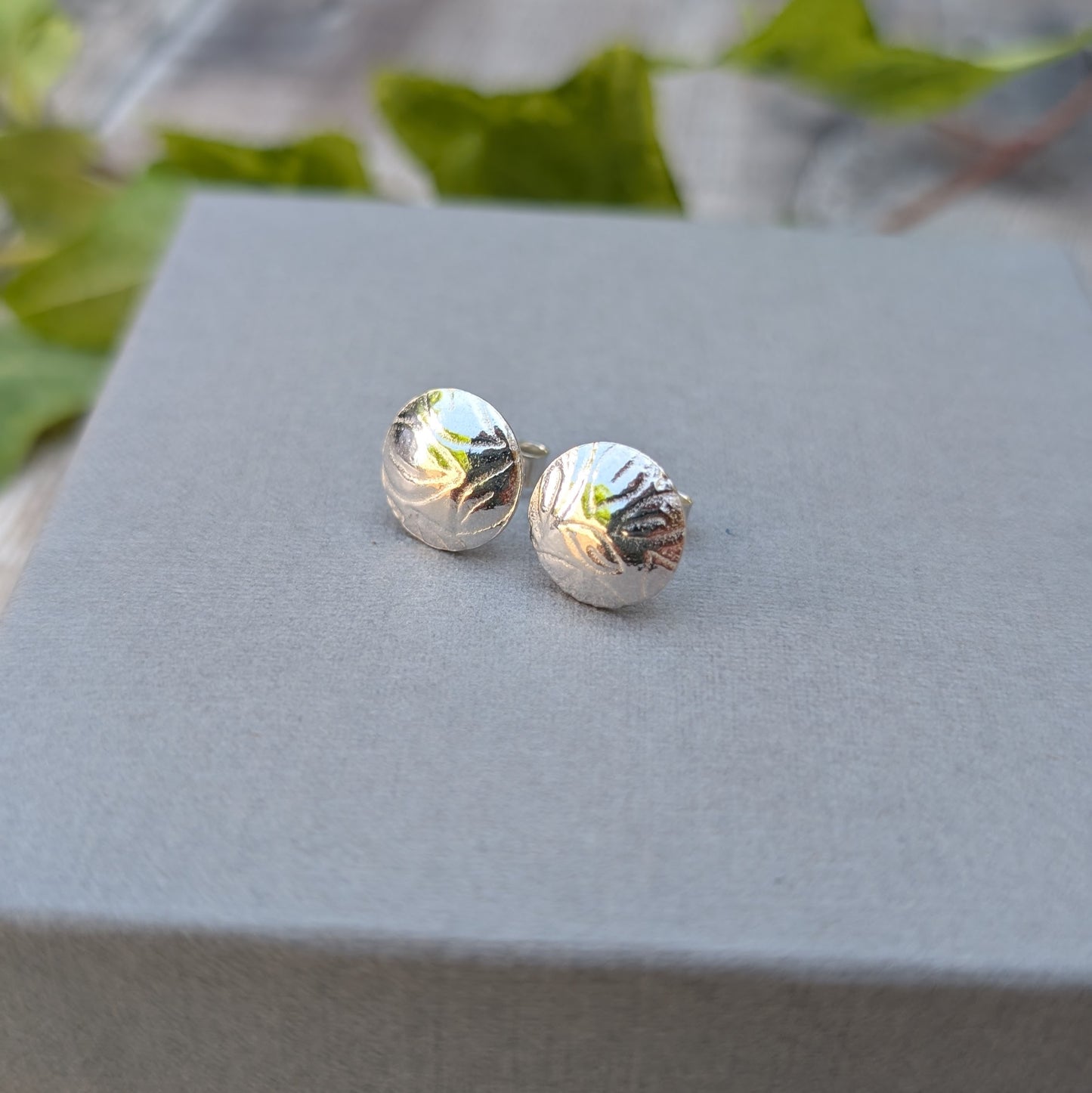 Silver Dome Disc Stud Earrings with Leaf Pattern displayed on a grey box with a soft outdoor background.