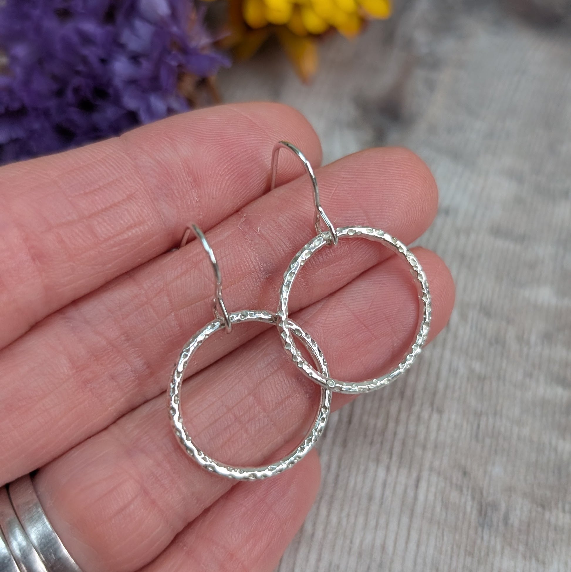 Hand holding a pair of Silver Dot Textured Circle Earrings, showing the hammered dot pattern on the circular drops with a blurred background of purple and yellow flowers.