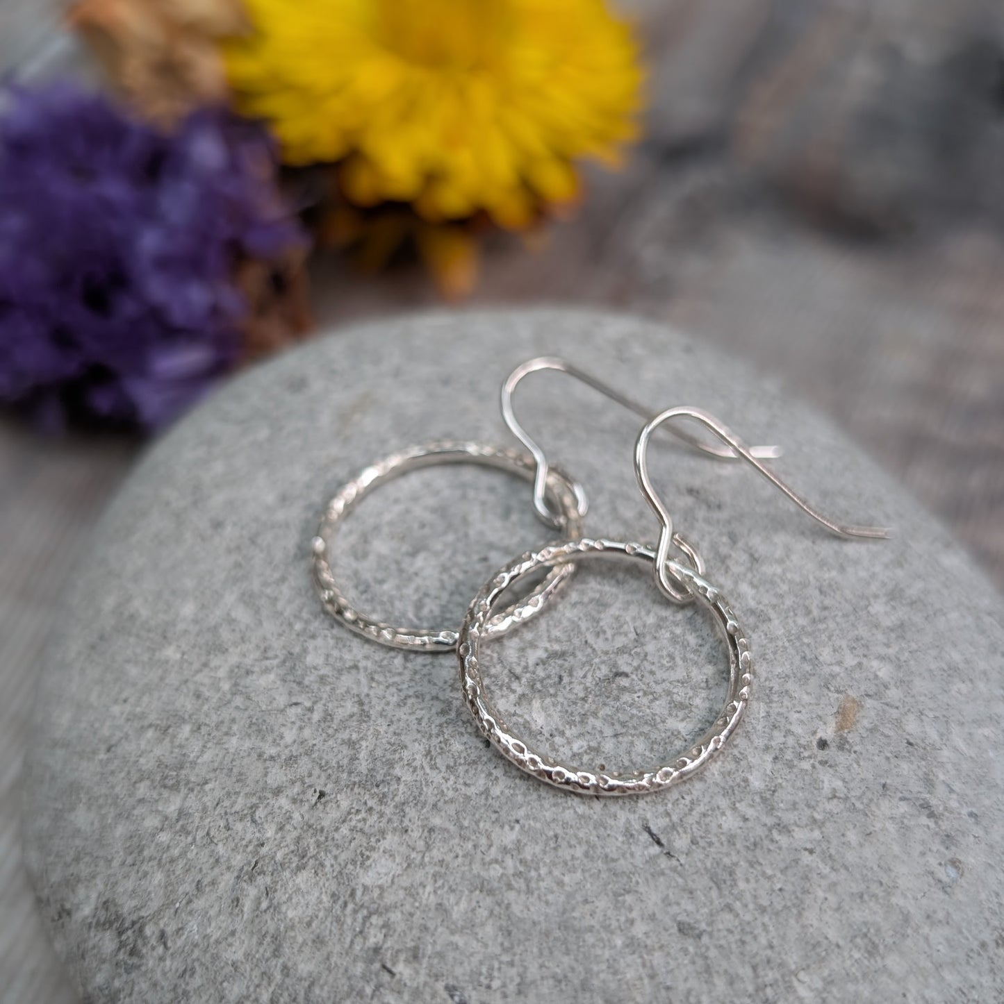 Silver Dot Textured Circle Earrings resting on a smooth grey stone, highlighting the fine dotted texture and simple hook ear wires.
