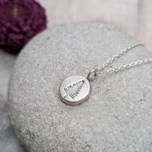 Another view of the silver evergreen tree pebble necklace on a grey stone with blurred flowers behind.