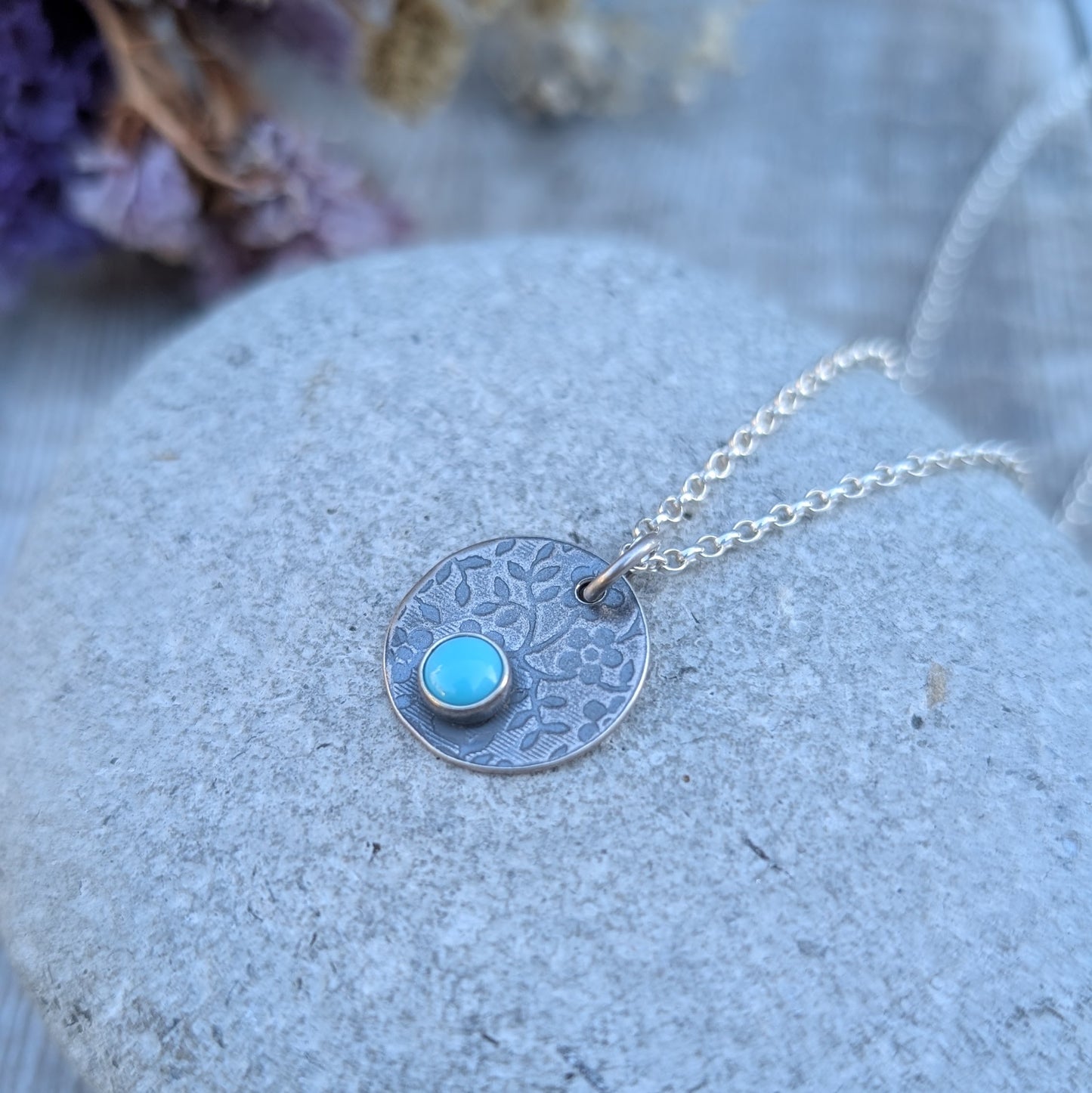 Close-up of silver floral disc necklace with Turquoise, resting on a stone with chain draped around it.