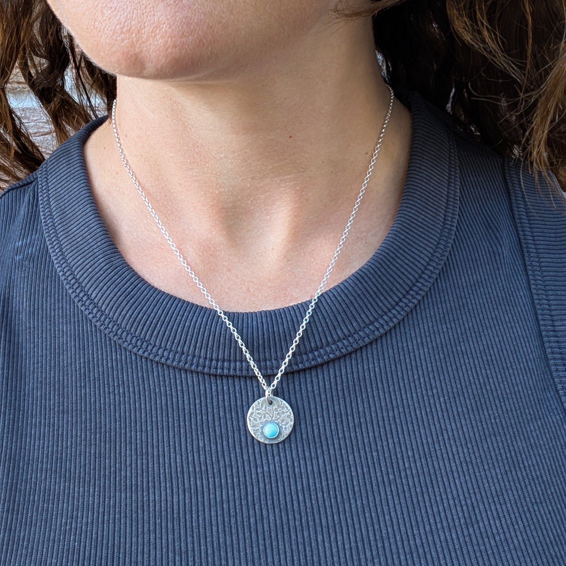 Silver floral disc necklace with Turquoise, worn on a silver chain against a dark top.