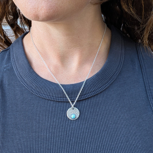Silver floral disc necklace with Turquoise, worn on a silver chain against a dark top.