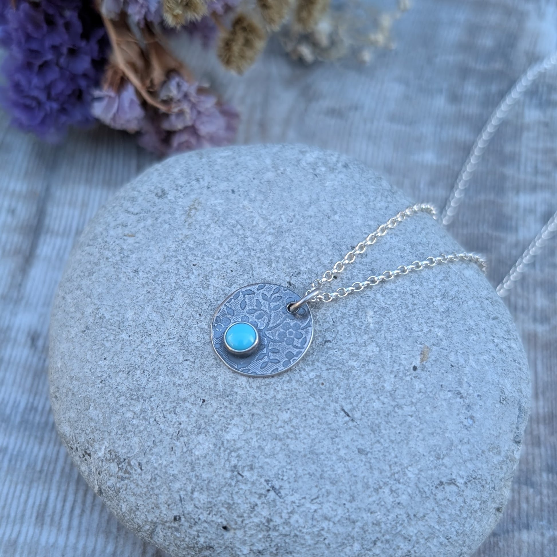 Silver floral disc necklace with Turquoise, worn on a silver chain against a dark top.