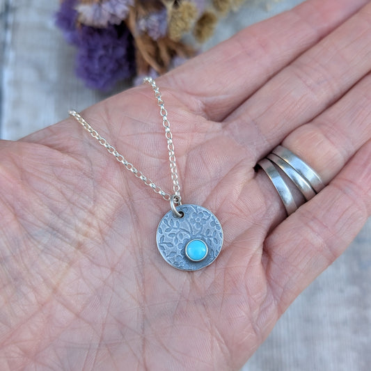 Hand holding silver floral disc necklace with Turquoise, showing the patterned detail of the pendant.