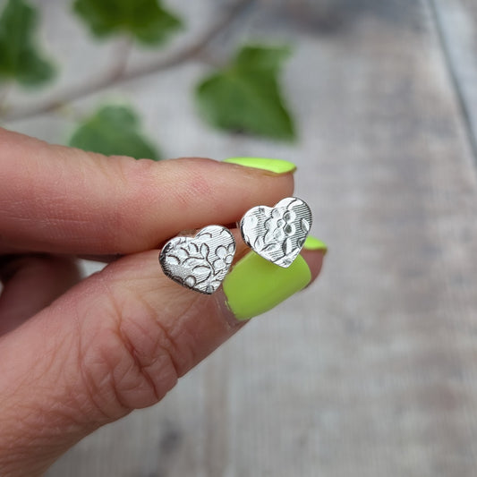Silver floral heart stud earrings held between fingers, showing the rolled floral pattern and small heart shape.