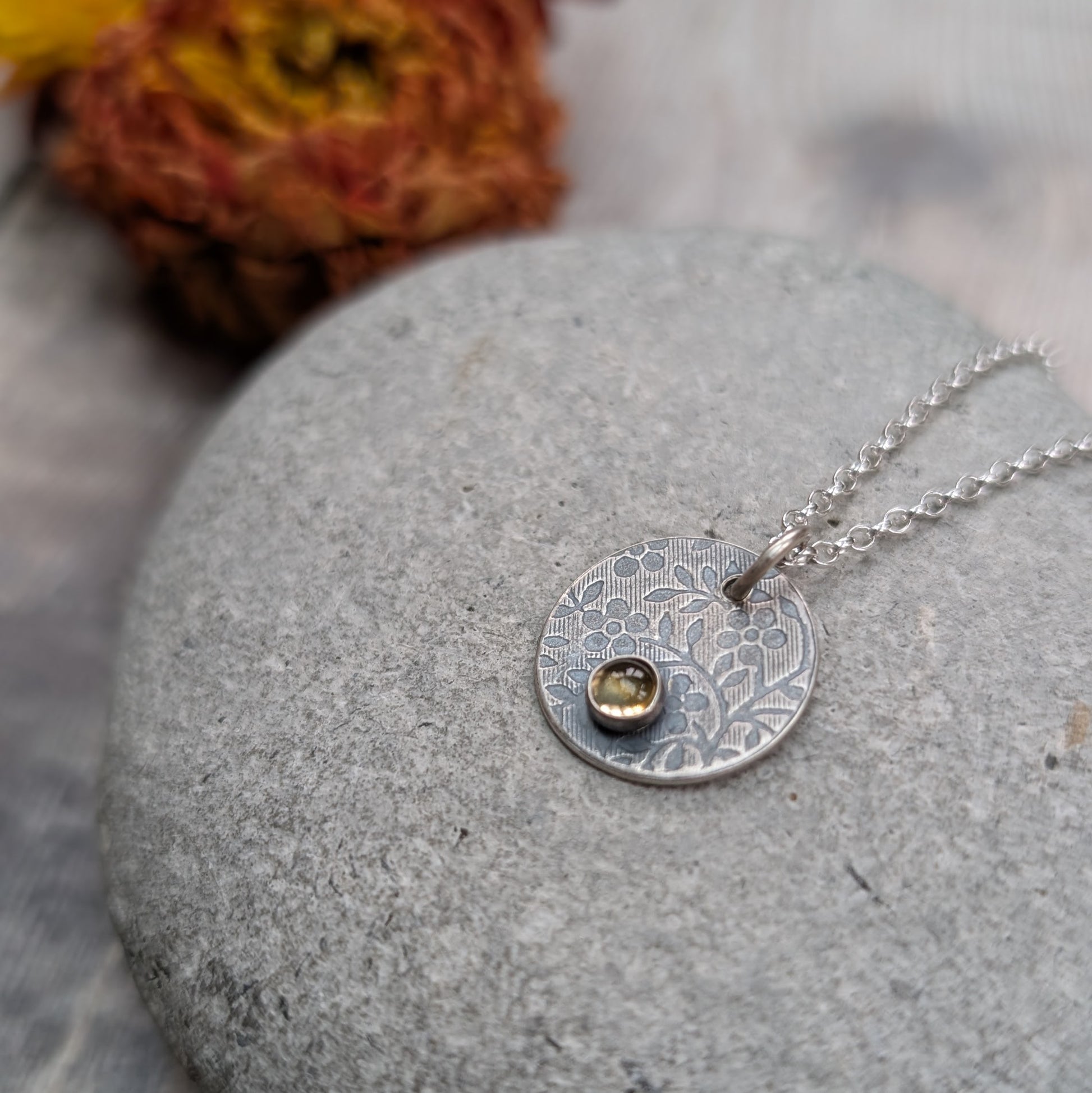 Silver flower disc necklace with citrine pendant resting on a stone, showing the detailed floral texture and yellow citrine gemstone.