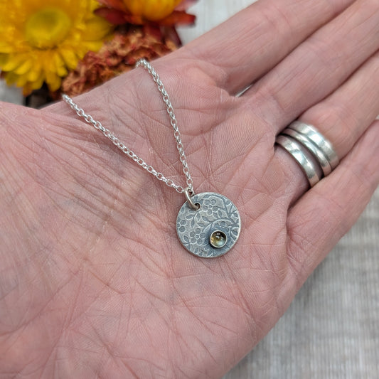 Silver flower disc necklace with citrine pendant displayed on an open hand, showing the patterned surface and gemstone detail.