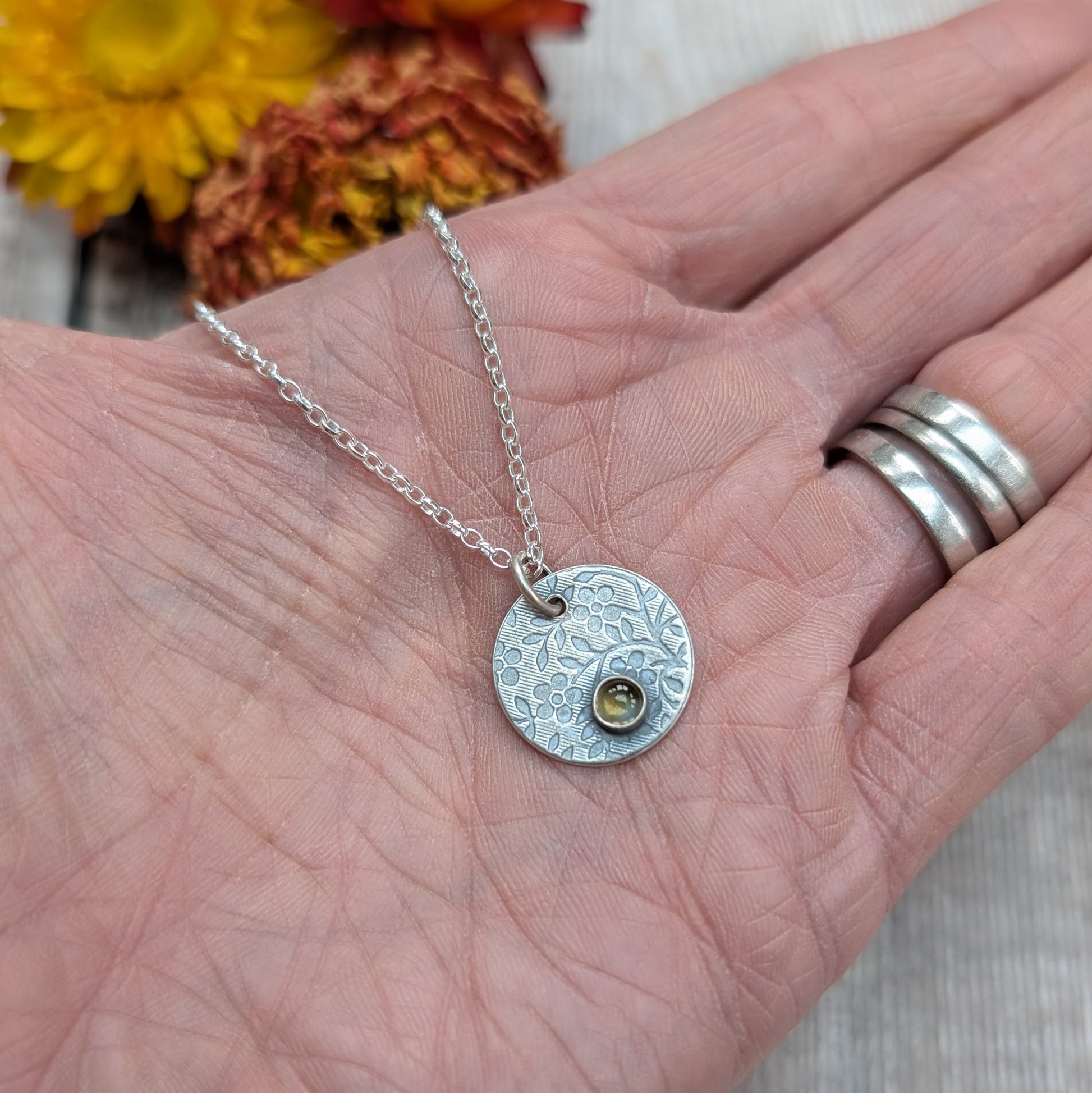Close-up of a silver flower disc necklace with citrine pendant in a hand, highlighting the floral pattern and golden gemstone.