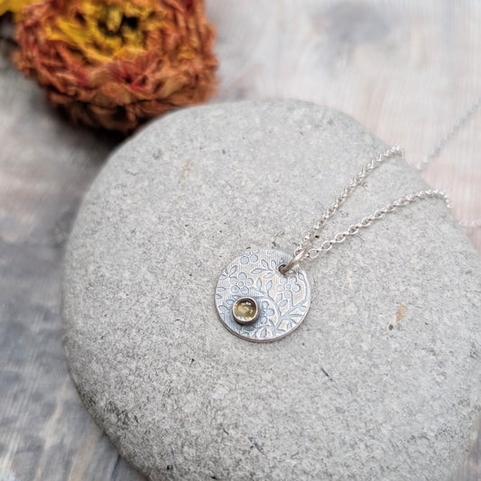 Silver flower disc necklace with citrine pendant resting on a smooth grey stone with orange flowers in the background.