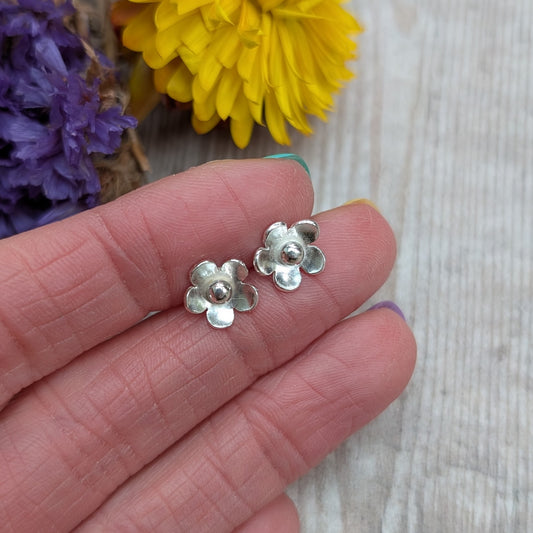 Close-up of a hand holding a pair of handmade silver flower stud earrings with a blurred yellow and purple flower background.