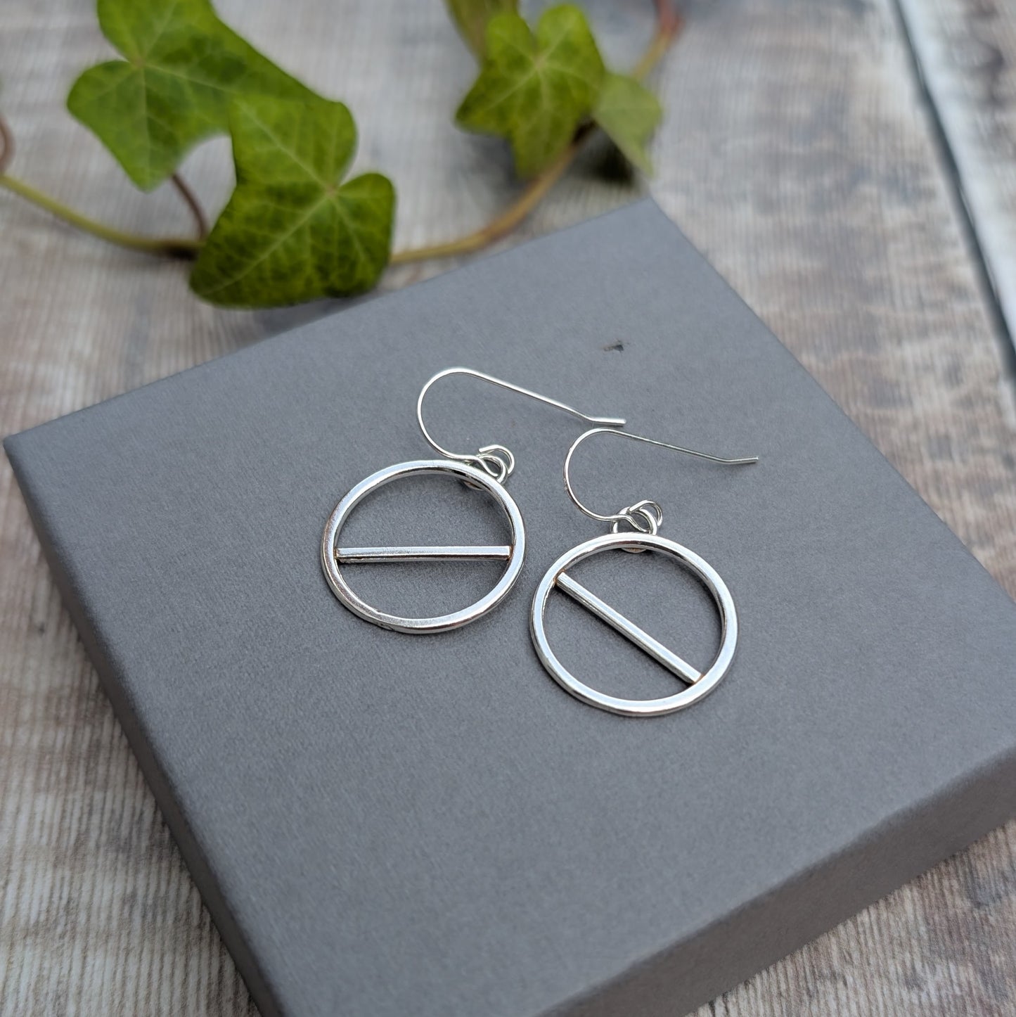 Silver geometric circle earrings handmade from recycled Sterling Silver, displayed on a grey jewellery box with green ivy in the background.