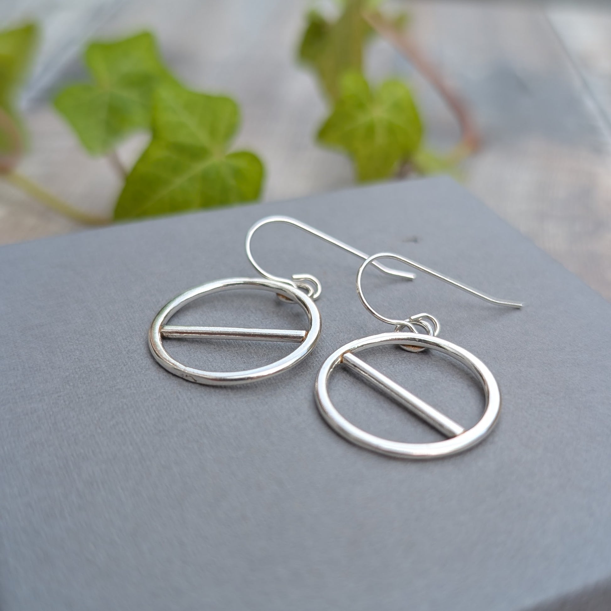Close up of silver geometric circle earrings with a simple straight bar design, resting on a grey jewellery box.