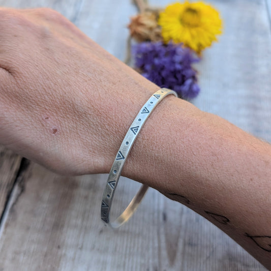 A silver geometric patterned bangle worn on a wrist, showing the hand-stamped triangle and dot design.